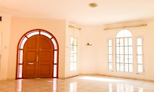 A spacious empty room with white tiled floor, large wooden front door, and tall arched windows allowing natural light to enter.