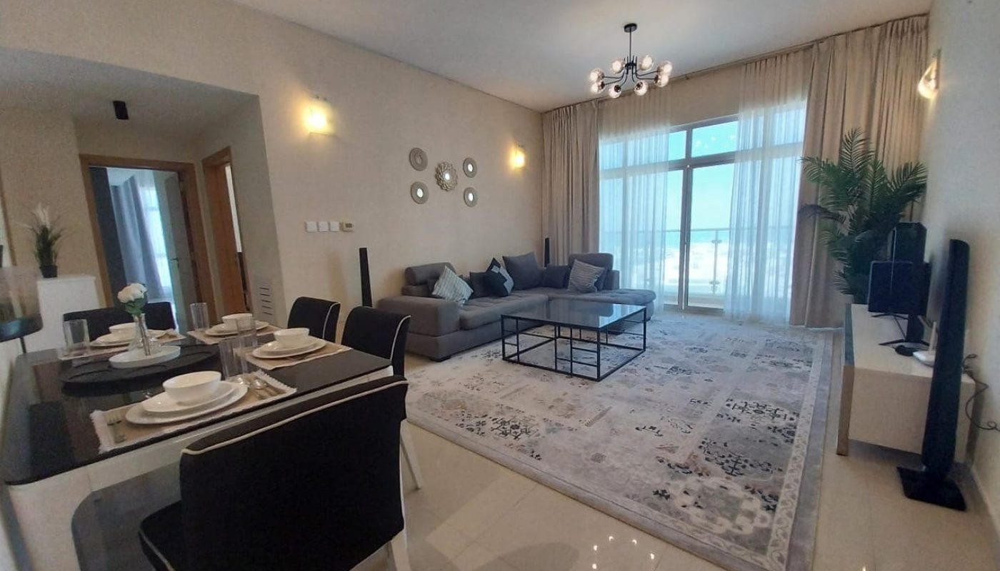 Modern living room and dining area with gray sofa, glass coffee table, flat-screen TV, dining table set for four, large windows with sheer curtains, and a potted plant.