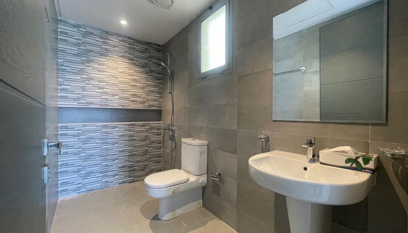 Modern bathroom with grey tiled walls, a wall-mounted sink, a toilet, a shower area, a mirror, and a small window providing natural light.