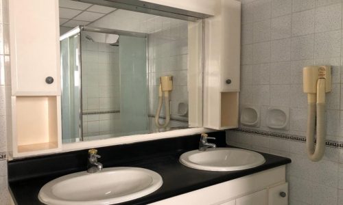 A bathroom with a double sink vanity, wall-mounted hairdryer, and a large mirror. The room features white tiles and a corner shower with glass doors.