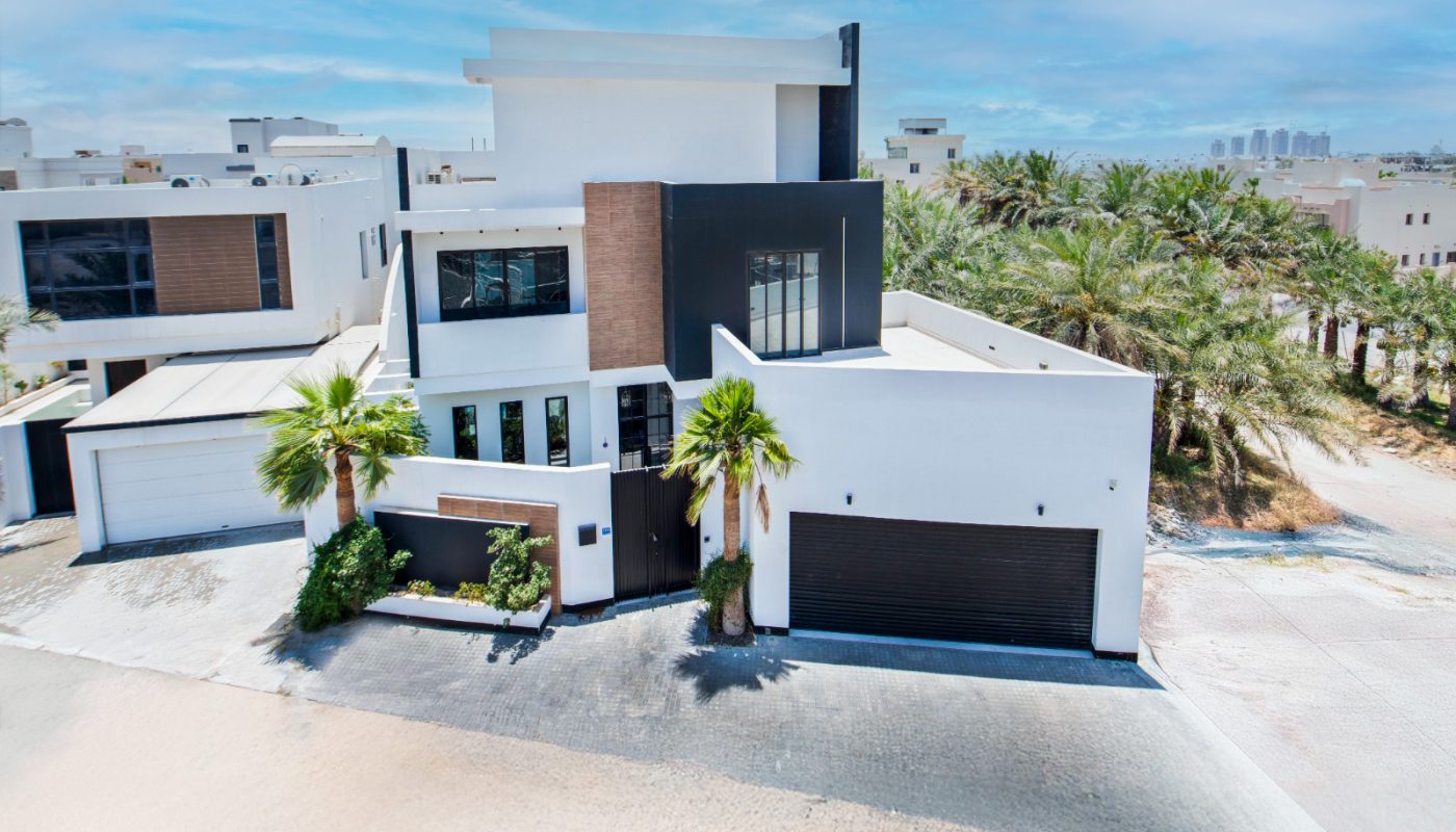 Modern three-story house with a black and white facade, garage, palm trees, and a paved driveway on a sunny day.