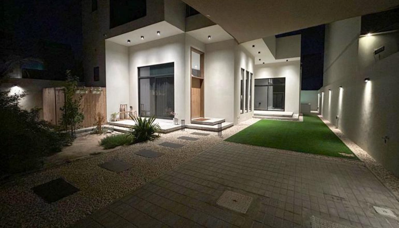 Modern house exterior at night with outdoor lights, large windows, a small lawn, and stone pathway. Minimalist landscaping with plants and gravel is visible.