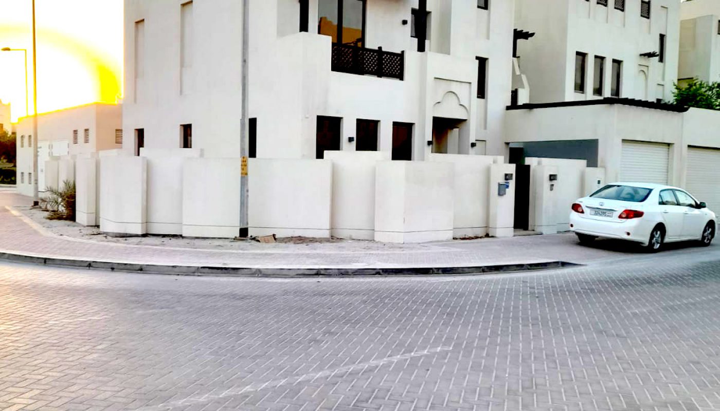 A white modern two-story house with balconies, a surrounding wall, and a white car parked outside on a paved street during sunset.