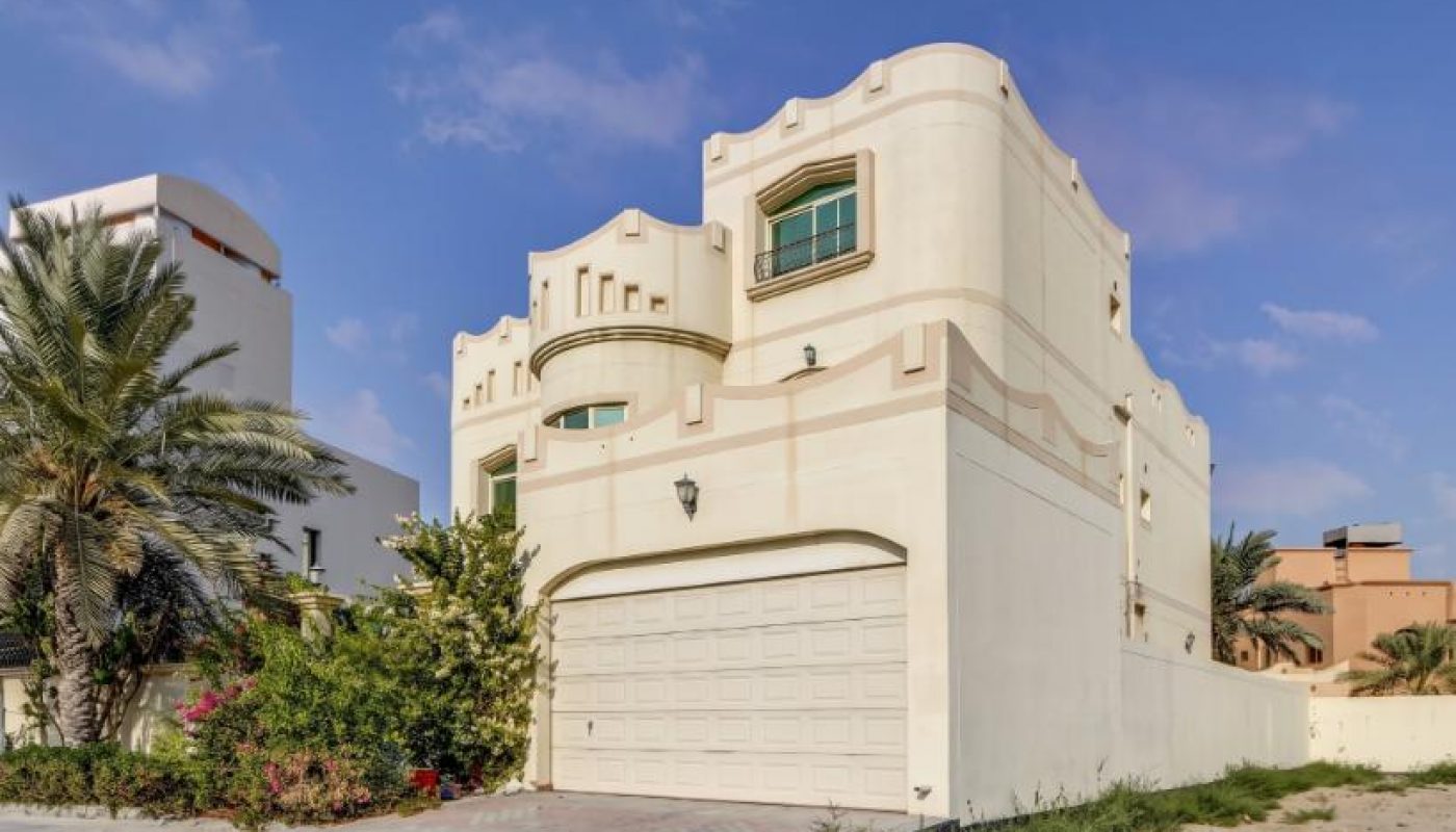 Two-story beige house with attached garage, arched windows, and minimal landscaping, set against a blue sky with some clouds.