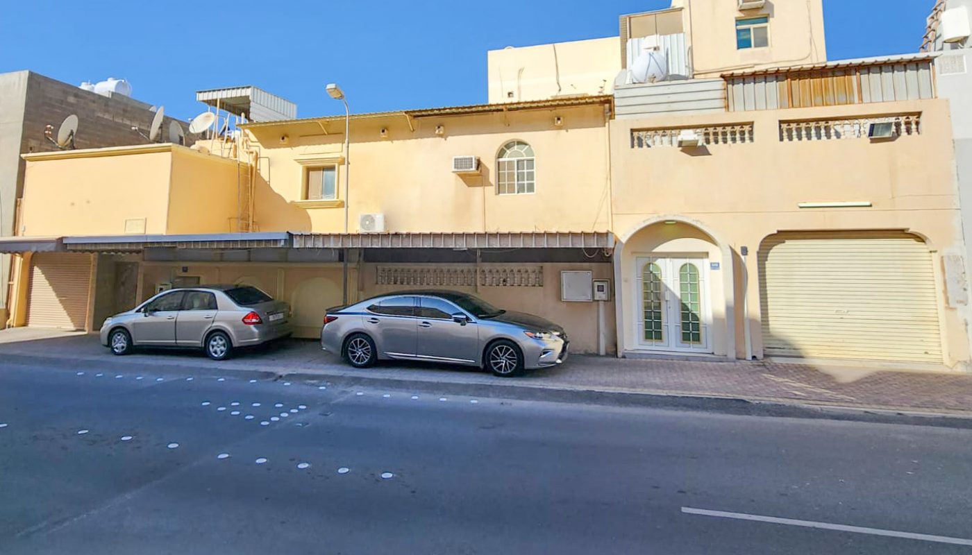 A beige residential building with multiple levels, two parked cars in front, and a clear blue sky overhead.