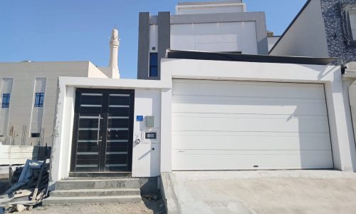 Contemporary 3BR Villa exterior under clear skies, featuring a garage door on the right and front door with sidelight on the left, alongside construction debris.