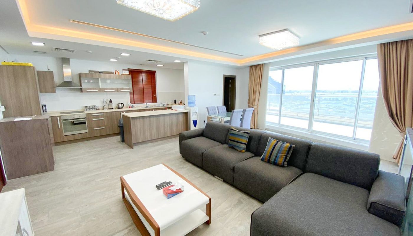 Modern living room with a large gray sectional sofa, a white coffee table, and an open kitchen with wooden cabinets. This luxury apartment in the Amwaj area features large windows with beige curtains offering an expansive view. Don't miss this opportunity; it's now for sale!