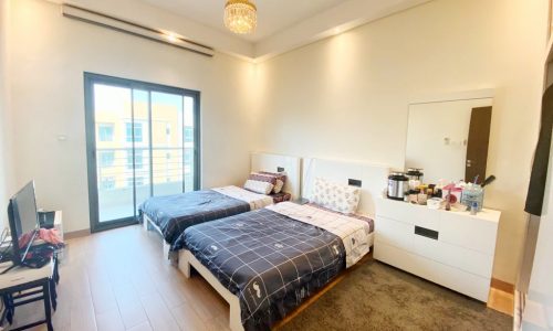 A bedroom with two twin beds, a wall-mounted TV, a white dresser with a mirror, and a sliding glass door leading to a balcony. A small chandelier hangs from the ceiling.