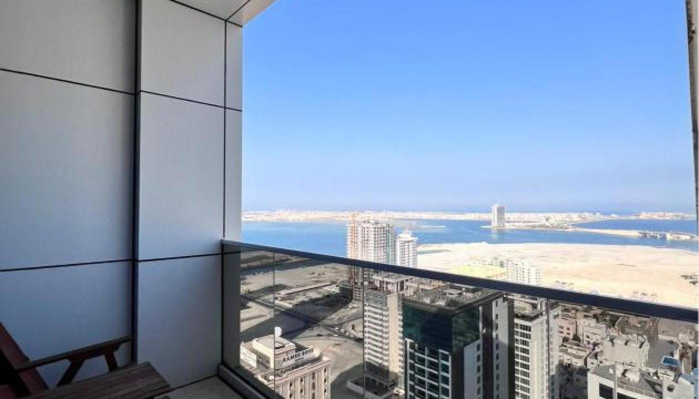 High-rise balcony with two wooden chairs overlooks city buildings and a sandy coastline under a clear blue sky.