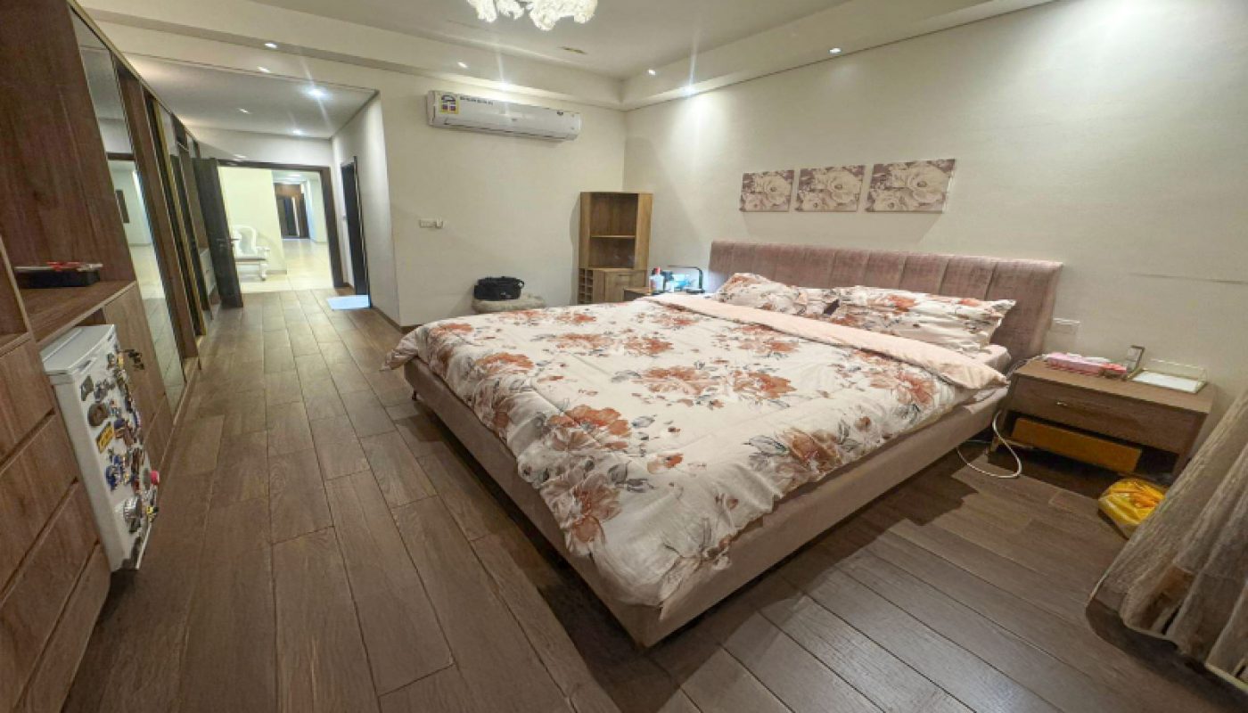 A spacious bedroom with a large bed, floral bedding, wooden floor, bedside tables, wardrobe, mini fridge, air conditioner, and wall art above the headboard.