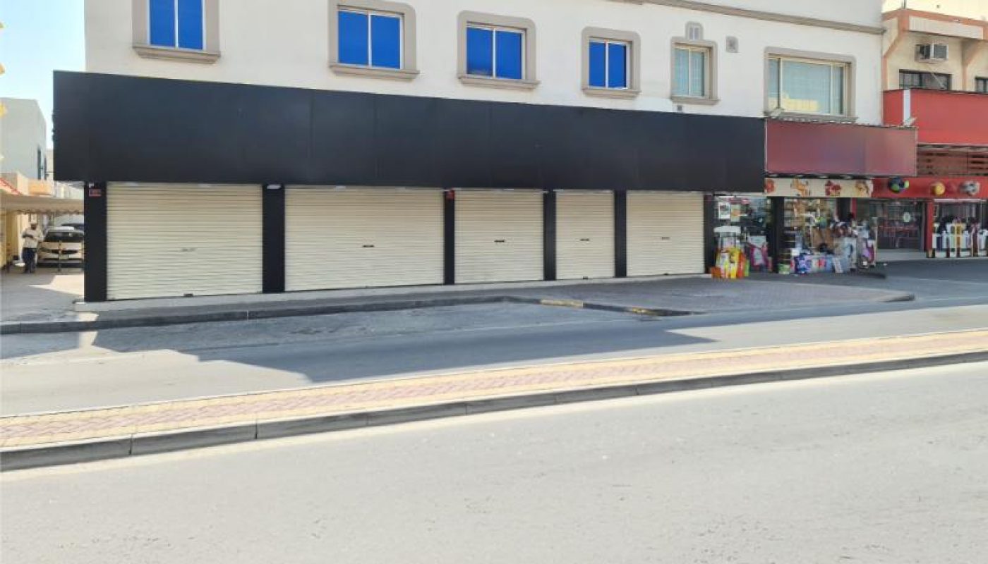 A two-story building with closed white shutters on the ground floor and blue-tinted windows above, situated on a quiet street with shops next door.