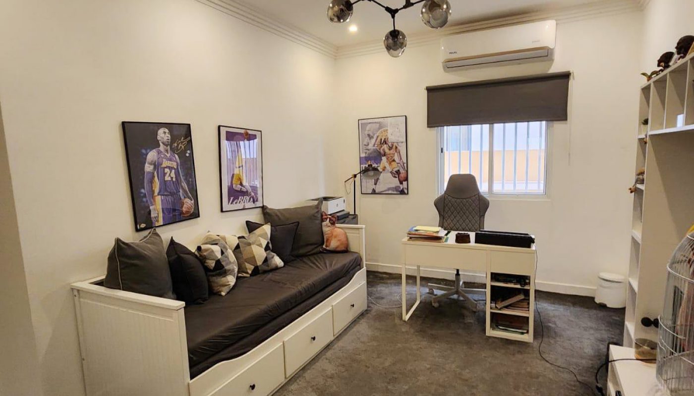 A modern home office with a couch, desk, chair, wall art, shelves, and an air conditioner under a ceiling light. The room has neutral colors and a window with a blind.