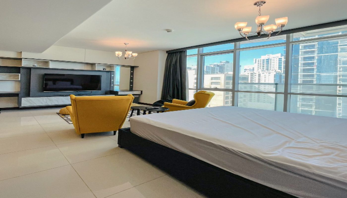 Modern studio apartment with large windows, a bed, yellow armchairs, a wall-mounted TV, shelving unit, and city view in the background.