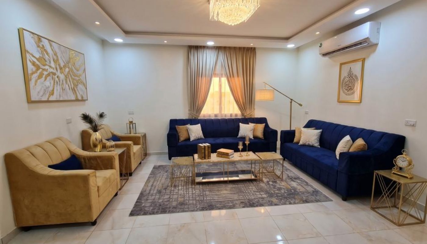 Modern living room with blue and gold furniture, abstract wall art, a chandelier, tile flooring, and a large window with sheer curtains letting in natural light.