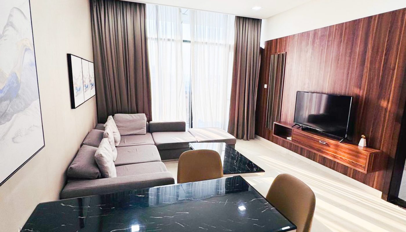 Modern living room with a gray sectional sofa, black marble dining table with brown chairs, wall-mounted TV, and large window with curtains letting in natural light.