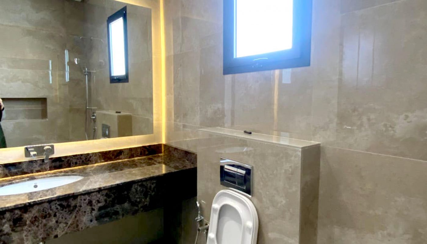 A modern bathroom with a wall-mounted toilet, a large mirror above a marble countertop sink, beige tiles, and a frosted window providing natural light.