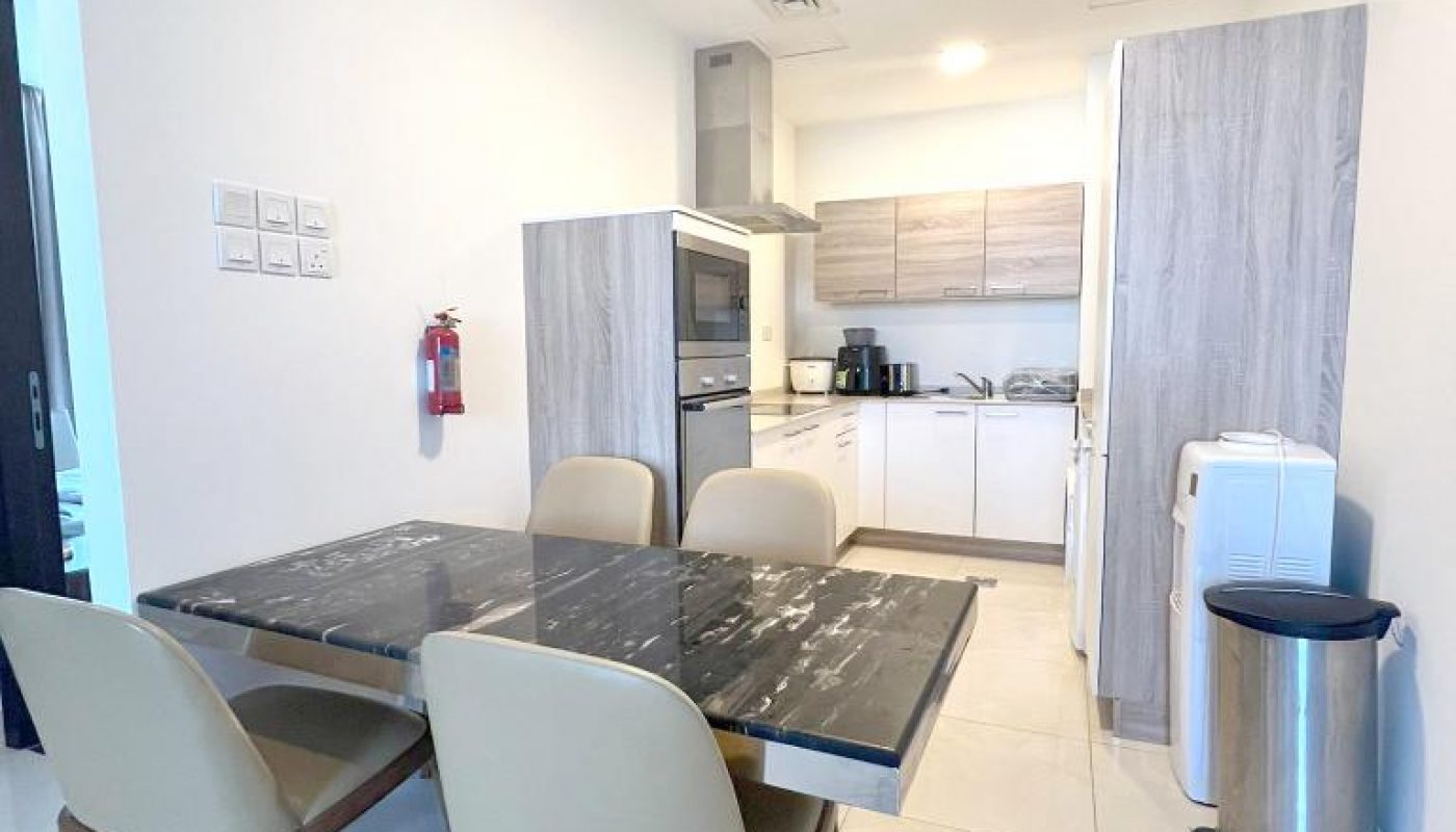 Modern kitchen and dining area with a black marble table, four beige chairs, light wood cabinets, appliances, a trash bin, and a fire extinguisher on a white tiled floor.