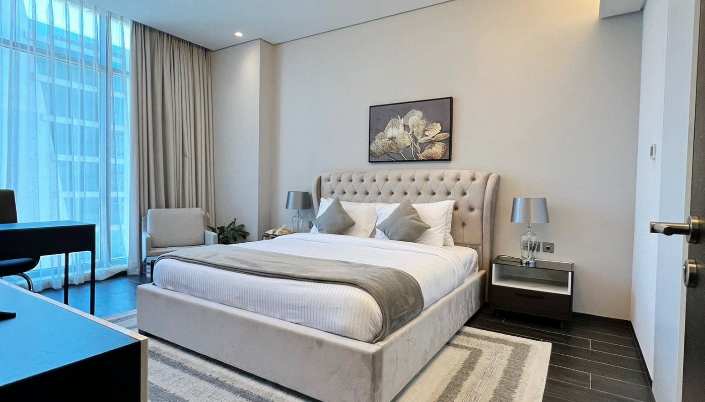 Modern bedroom with a large bed, two bedside tables with lamps, a desk by the window, and a patterned rug on dark wood flooring. Light streams in through floor-to-ceiling windows.