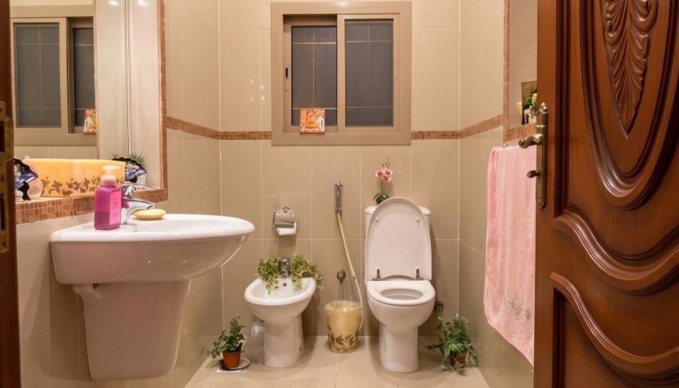 A bathroom with a sink, bidet, toilet, a large decorative rug, wall mirror, small plants, and beige tiled walls.