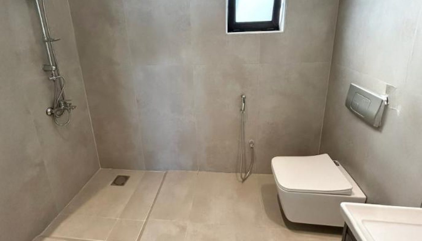 A modern bathroom with a walk-in shower, wall-mounted toilet, bidet spray, small window, and a sink with a dark cabinet.
