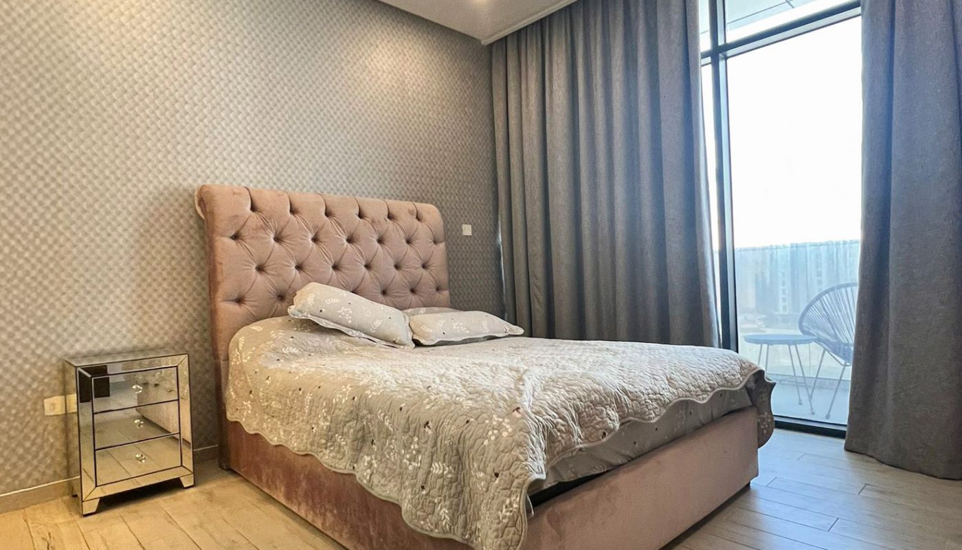 Modern bedroom with a tufted bed, mirrored nightstand, light wood floor, gray walls, and large window with floor-to-ceiling curtains leading to a balcony.