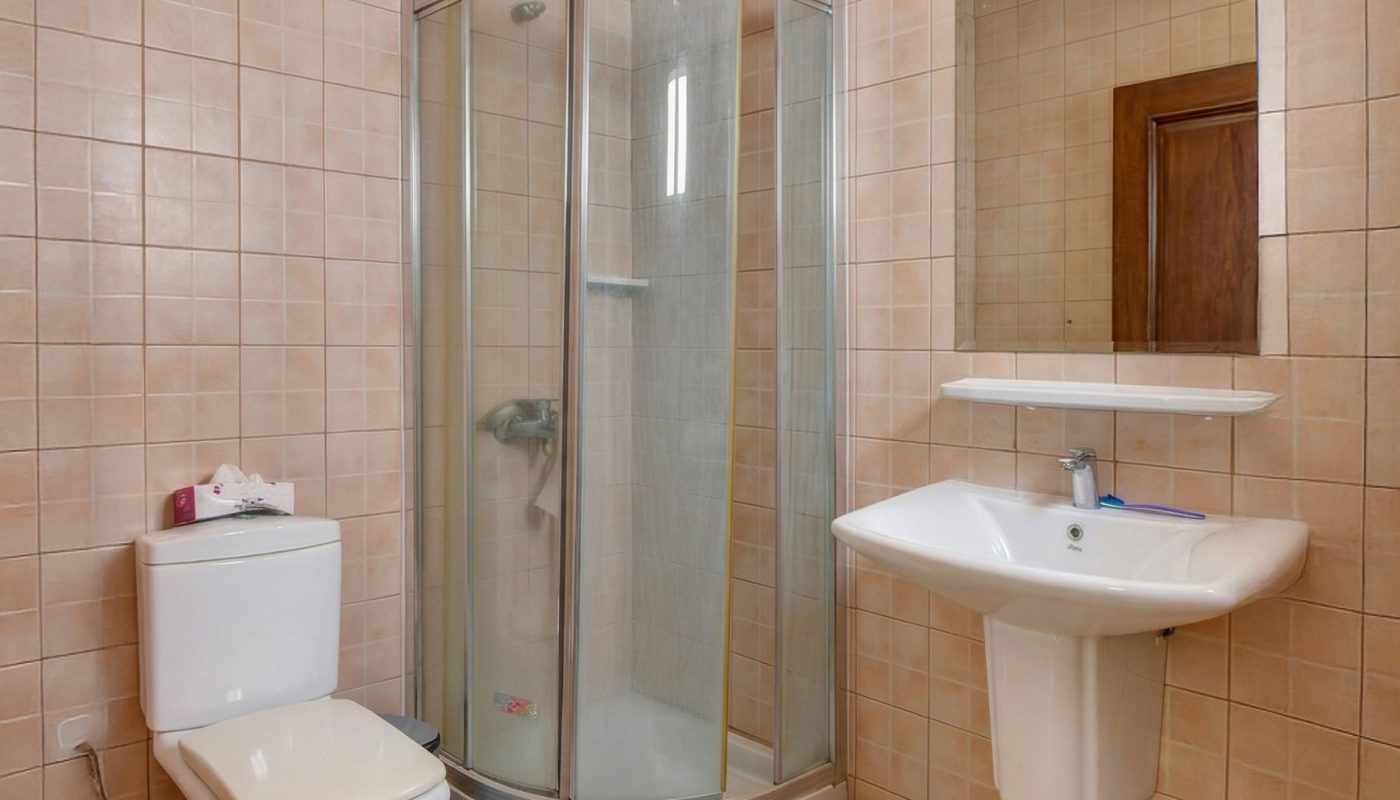 Bathroom with beige tiled walls and floor, featuring a glass-enclosed corner shower, a white toilet, a wall-mounted sink with a mirror, and a small shelf above the sink.