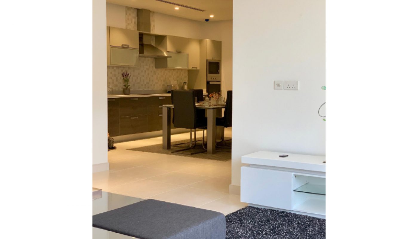 Modern apartment interior showing an open-plan kitchen and dining area with neutral colors, viewed from a living room with dark furnishings and a light-colored floor.
