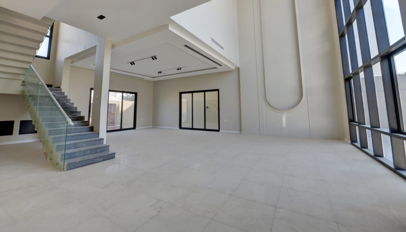 Spacious modern interior with high ceilings, large floor-to-ceiling windows, tiled floor, and a glass-railed staircase leading to an upper level.