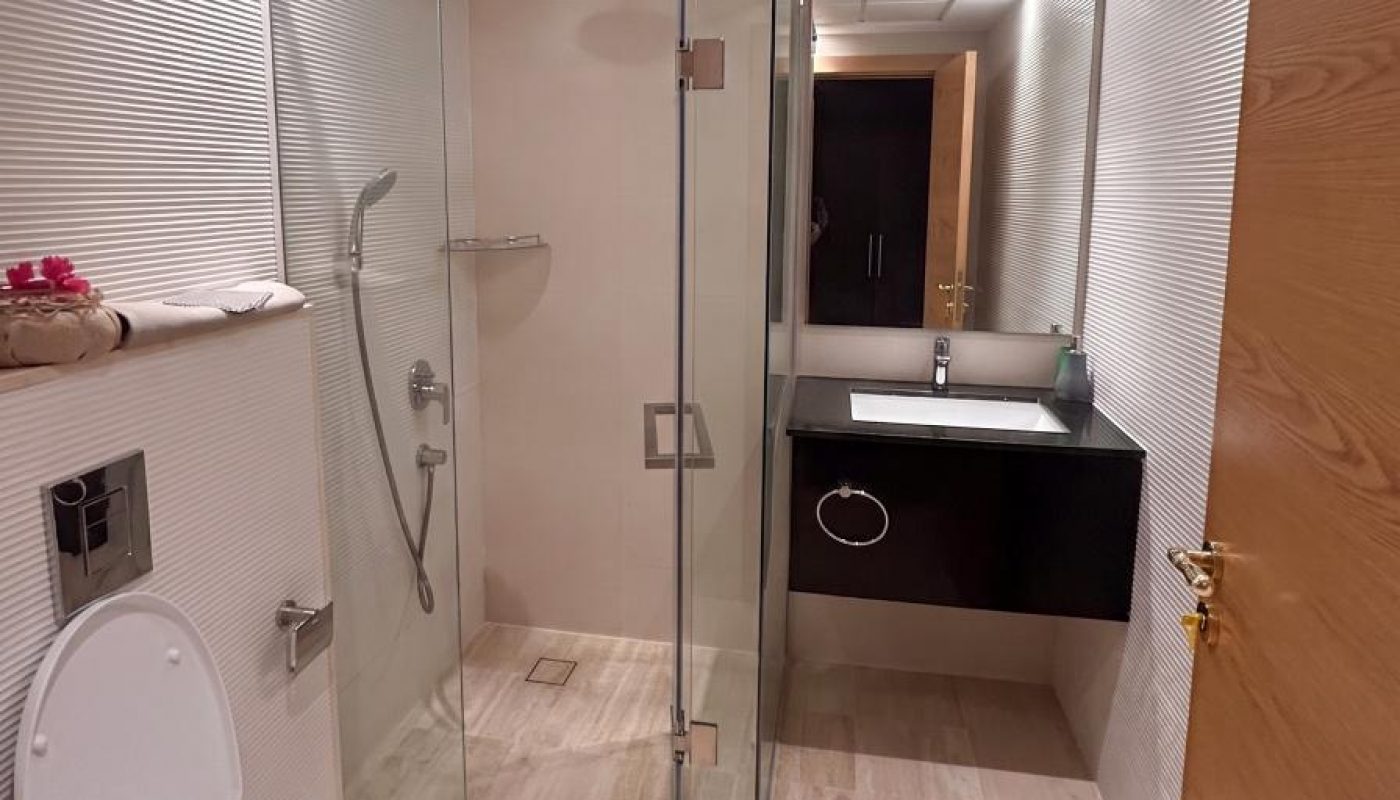 Modern bathroom with an open toilet, glass-enclosed shower, wall-mounted sink with mirror, and light-colored tile flooring and walls.