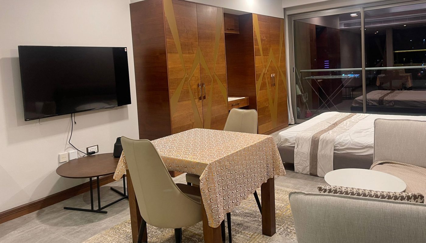 A modern studio apartment with a dining table, chairs, wall-mounted TV, bed, armchair, and a balcony with a nighttime city view.