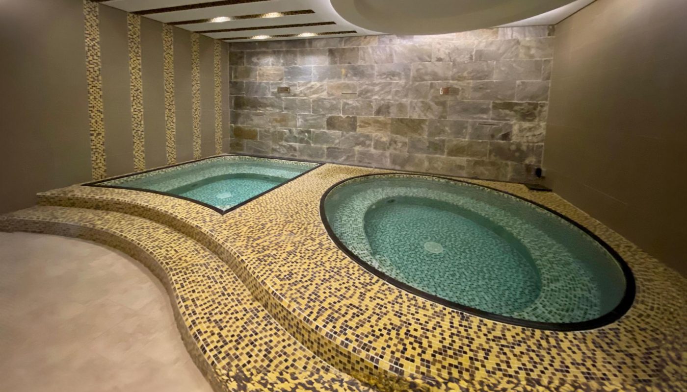 Two round, mosaic-tiled hot tubs are set in a modern indoor spa room with stone walls and curved ceiling lighting.