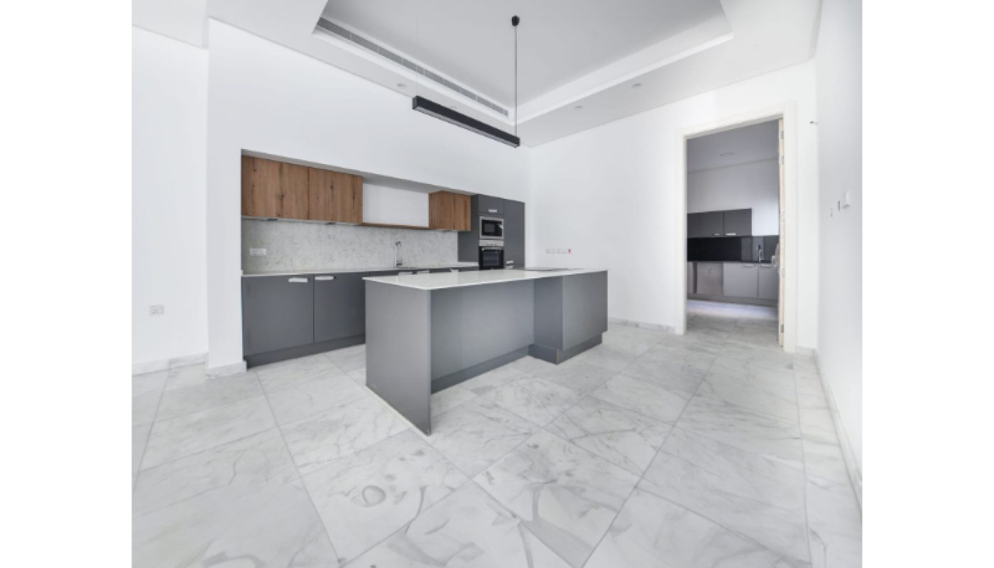 Modern kitchen with a central island, marble floors, grey and wood cabinets, white walls, and a black pendant light. An open doorway leads to another room in the background.