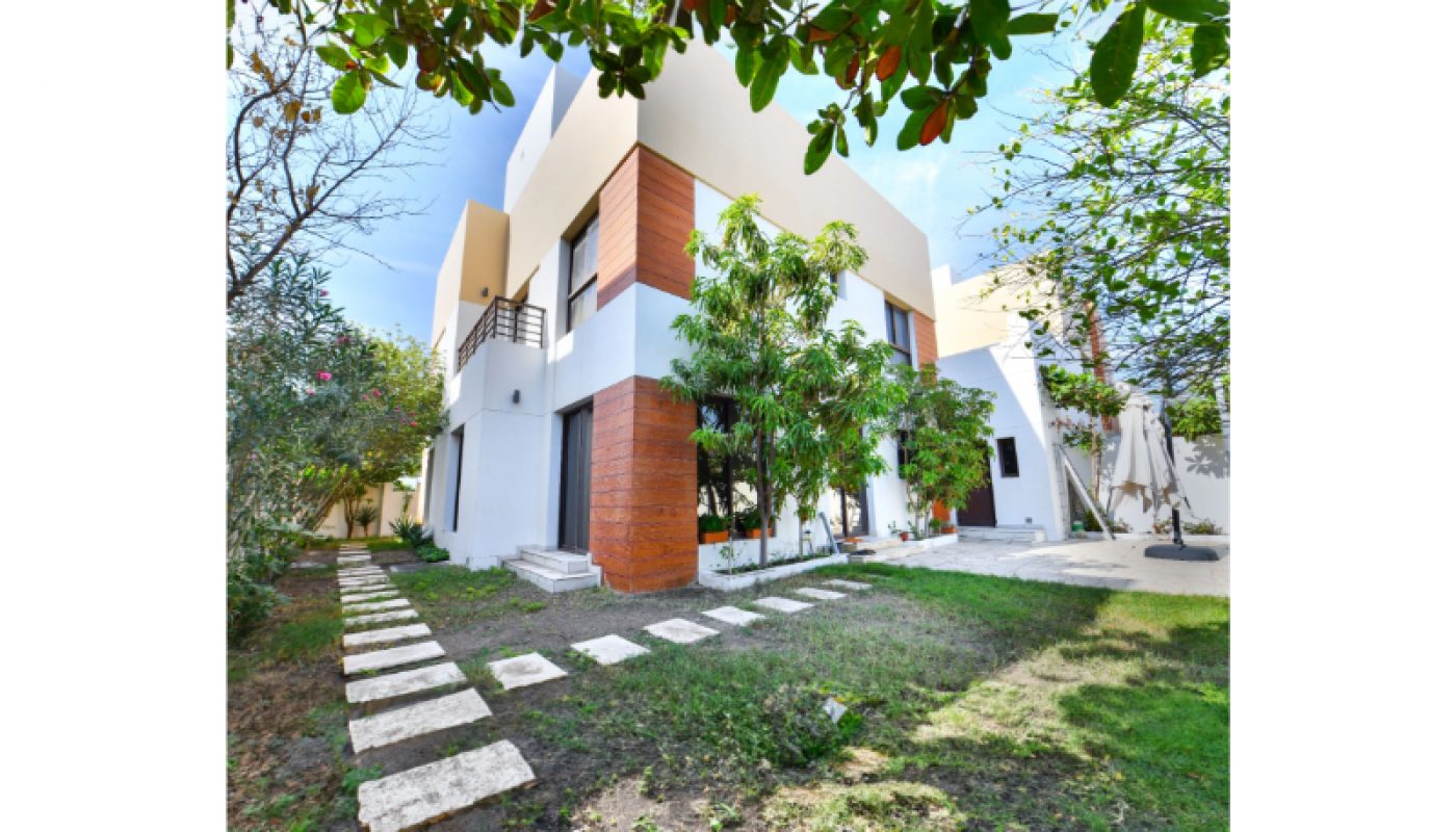 Modern two-story house with white walls and brown accents, surrounded by trees and a garden with stone stepping paths.