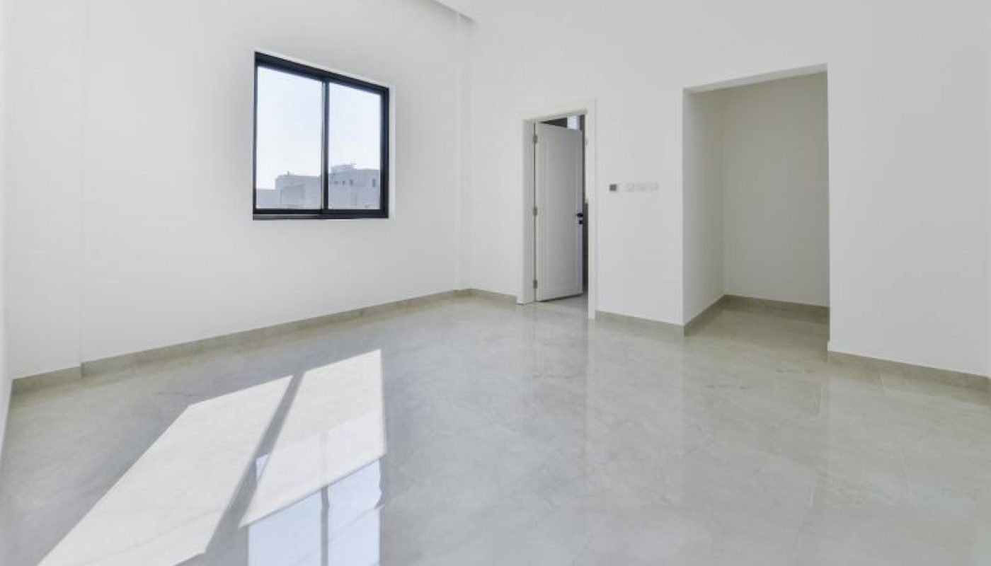 A spacious, empty room with white walls, a tiled floor, one large window, and two doorways. Sunlight enters through the window, casting shadows on the floor.