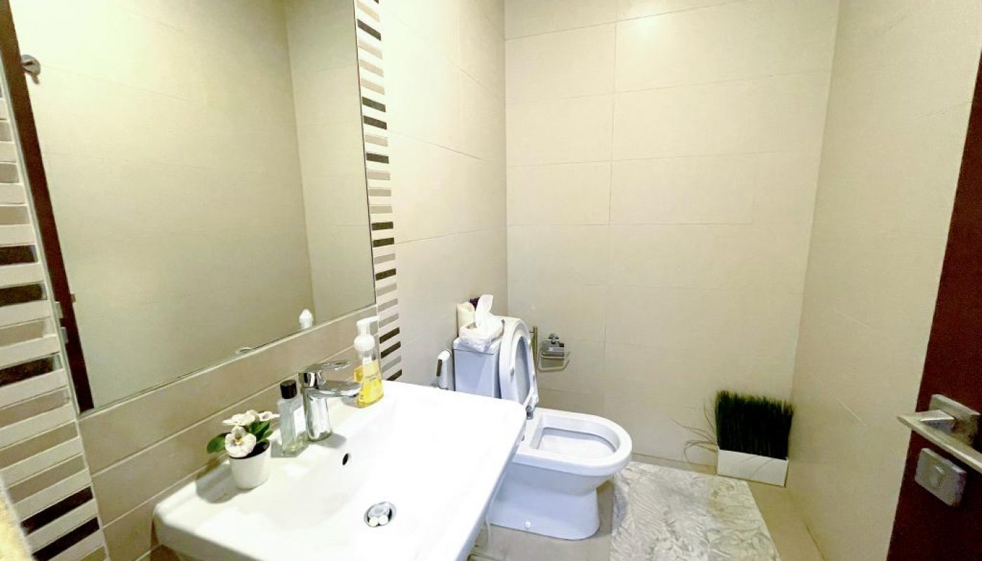 Modern bathroom with a large mirror, white sink, soap dispensers, toilet, bidet, a decorative plant, and beige tiled walls and floor.