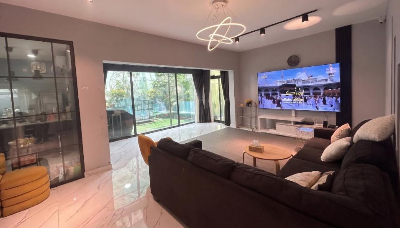 Modern living room with dark sectional sofa, large TV showing Mecca, and glass display cabinet.