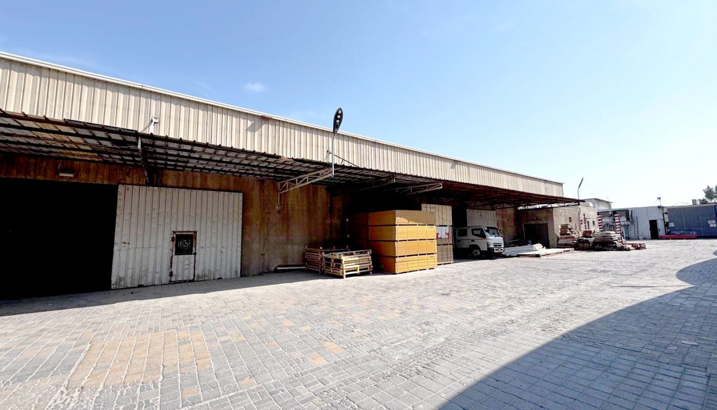 A warehouse with a metal exterior, stacked pallets, a white truck, and construction materials in an open paved yard under a clear sky.