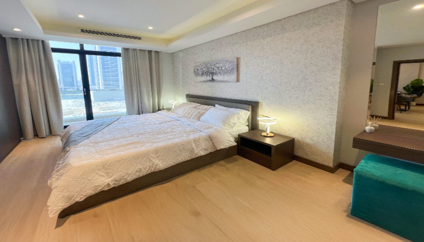 Modern bedroom with a large bed, two bedside tables with lamps, a wall mirror, and floor-to-ceiling windows overlooking city buildings.