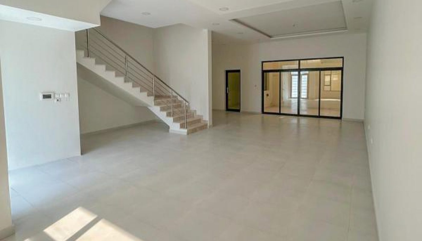 Spacious, empty modern room with large windows, tiled floor, staircase to the left, and glass doors at the far end.