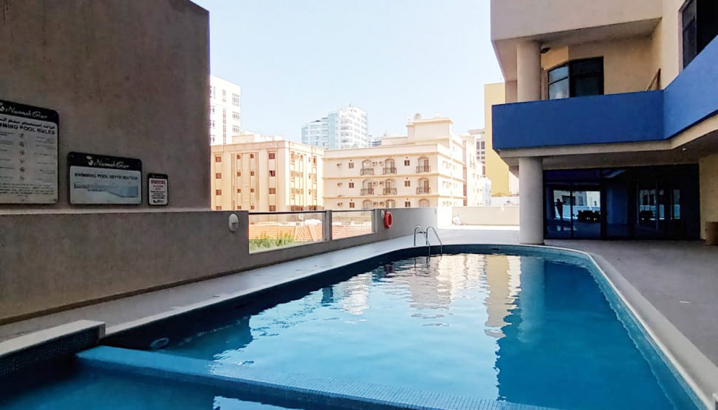 Outdoor swimming pool next to a modern apartment building, with city buildings in the background and safety signs on the wall.
