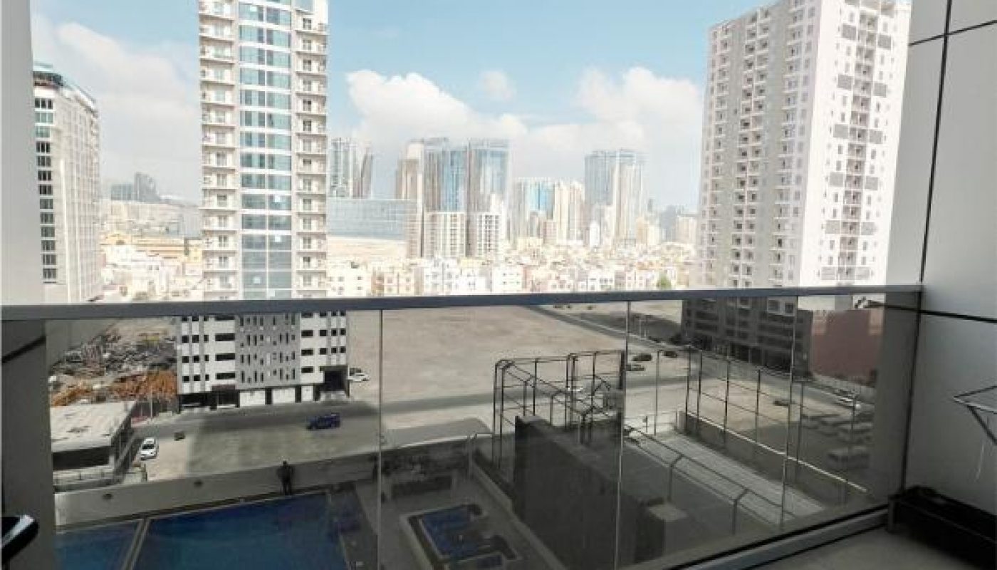 High-rise city view from a tiled balcony with glass railing, overlooking a swimming pool and surrounding tall buildings under a partly cloudy sky.