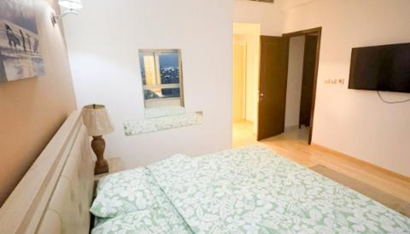 A bedroom with a double bed, floral bedding, a wall-mounted TV, a mirror, a lamp, and overhead lighting.
