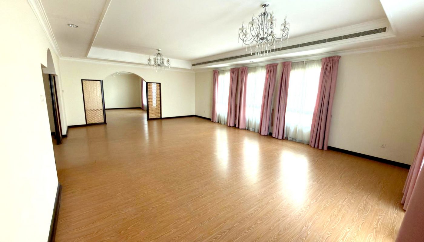 Spacious empty room with light wood flooring, cream walls, pink curtains, chandeliers, and an arched doorway leading to another room.
