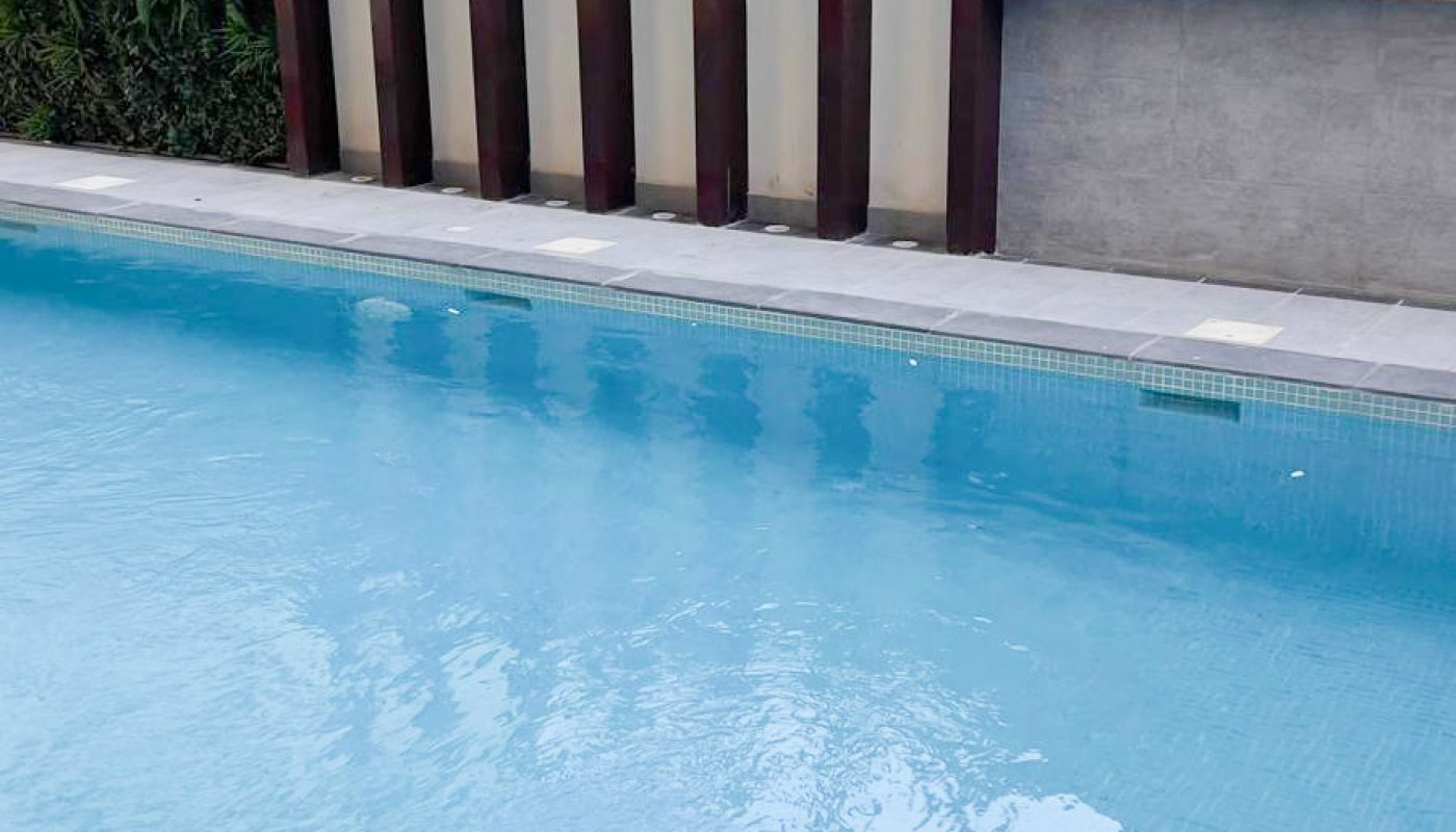 A rectangular outdoor swimming pool with clear blue water next to modern buildings and a fence with vertical wooden slats.