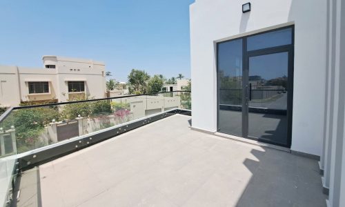 A spacious balcony with a glass railing overlooking a peaceful residential area, featuring large black-framed sliding doors and a clear blue sky.