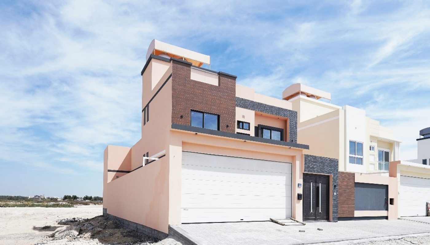 A modern two-story house with a flat roof, large white garage door, and brown and beige exterior walls, situated on a sandy lot under a blue sky with scattered clouds.