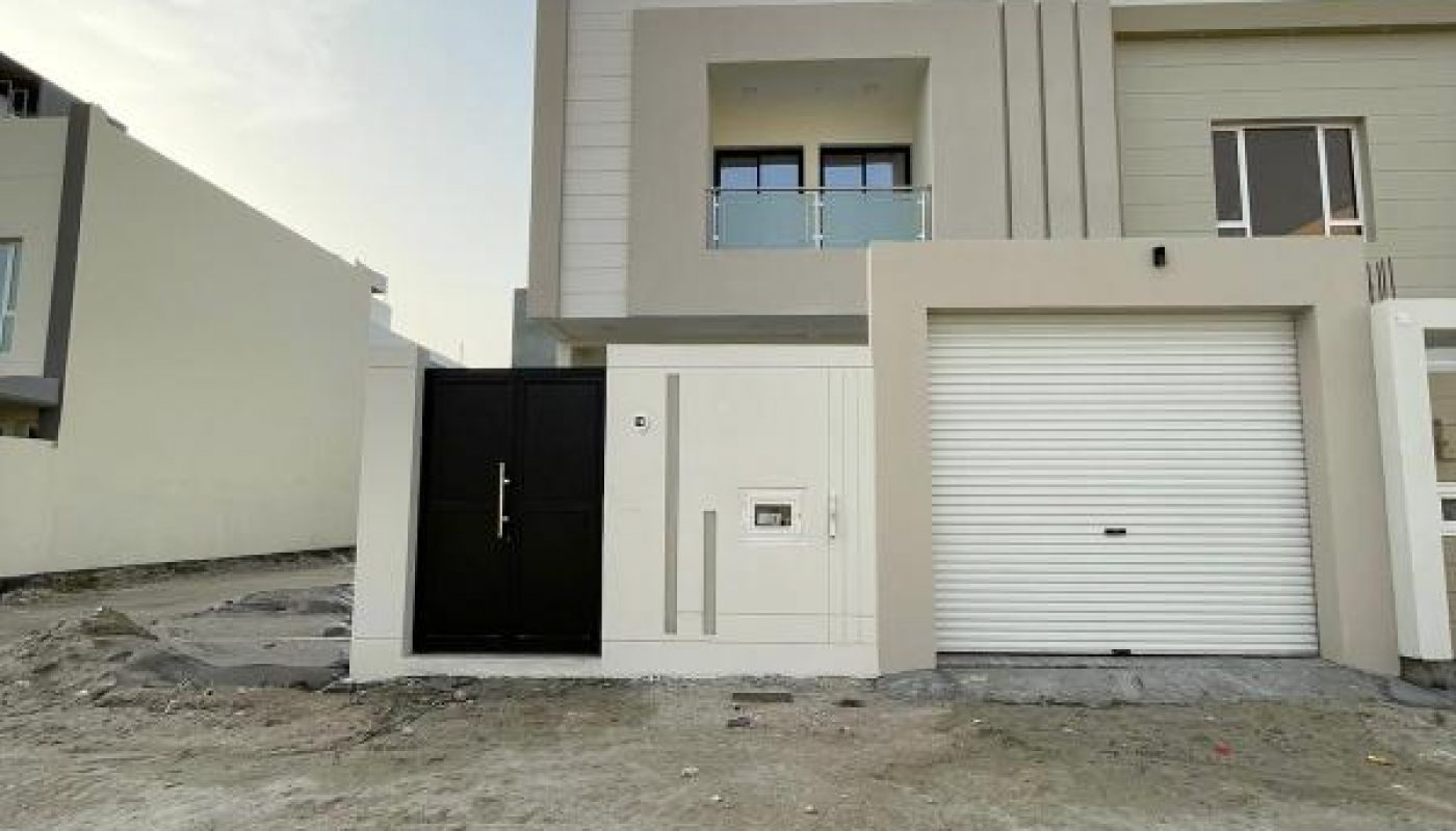 Two-story modern house with a balcony, black entrance door, white garage door, and unfinished front yard.
