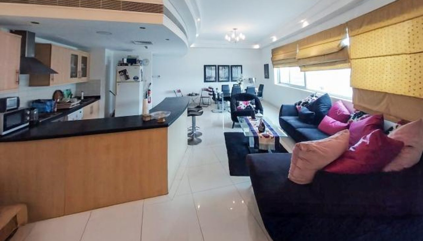 Modern open-plan kitchen and living room with black sofas, colorful cushions, tiled floor, and large windows with yellow blinds.