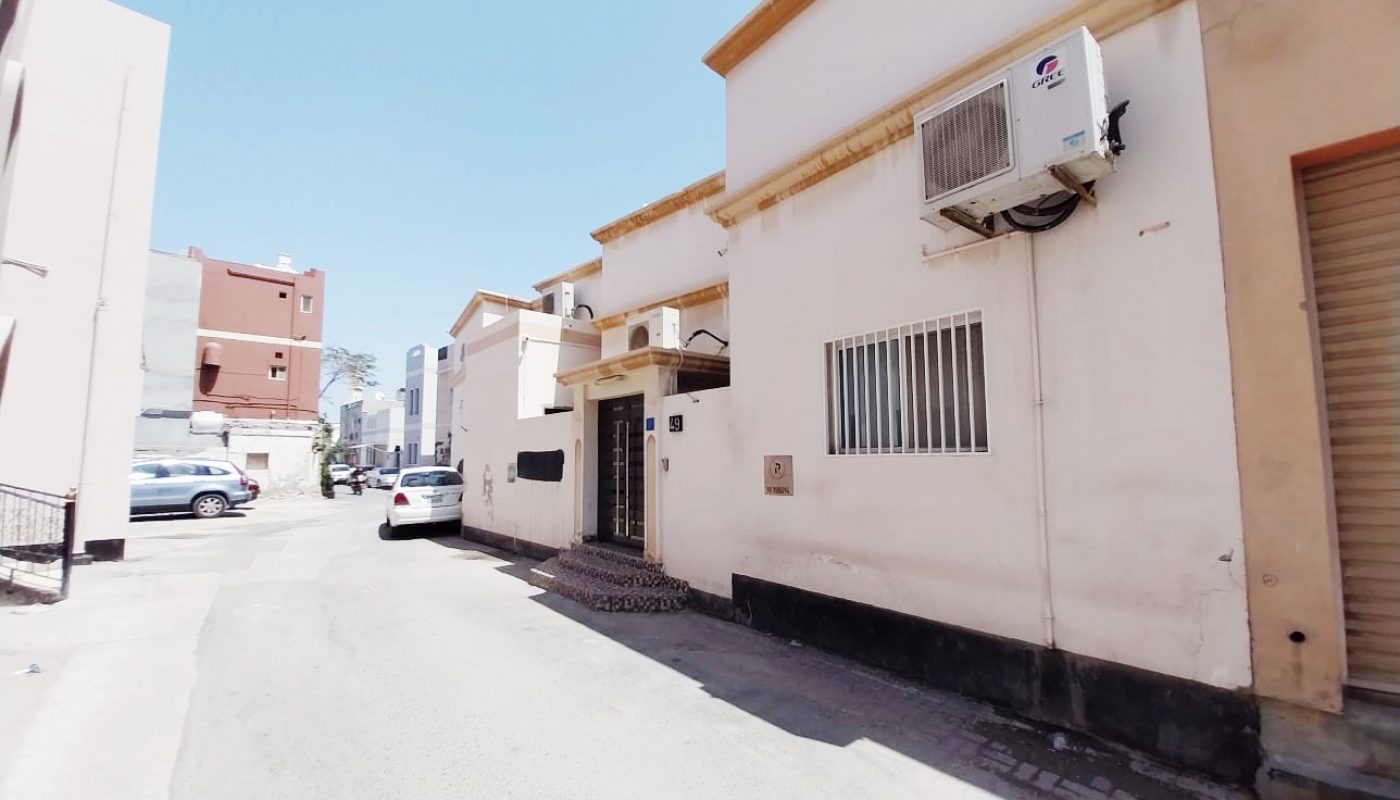 A narrow street with white buildings, a wall-mounted air conditioner, barred windows, a closed garage door, and a white car parked in the background under a clear sky.