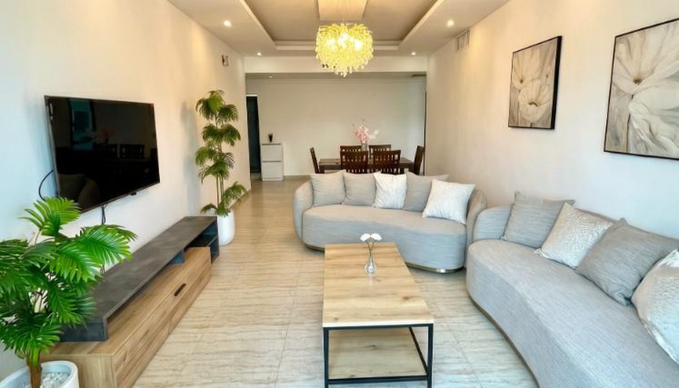 Modern living room with a curved gray sofa, wooden coffee table, wall-mounted TV, potted plants, and framed art on white walls; dining area visible in the background.